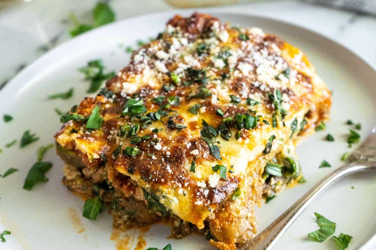 A slice of baked lasagna topped with melted cheese and fresh herbs on a white plate.