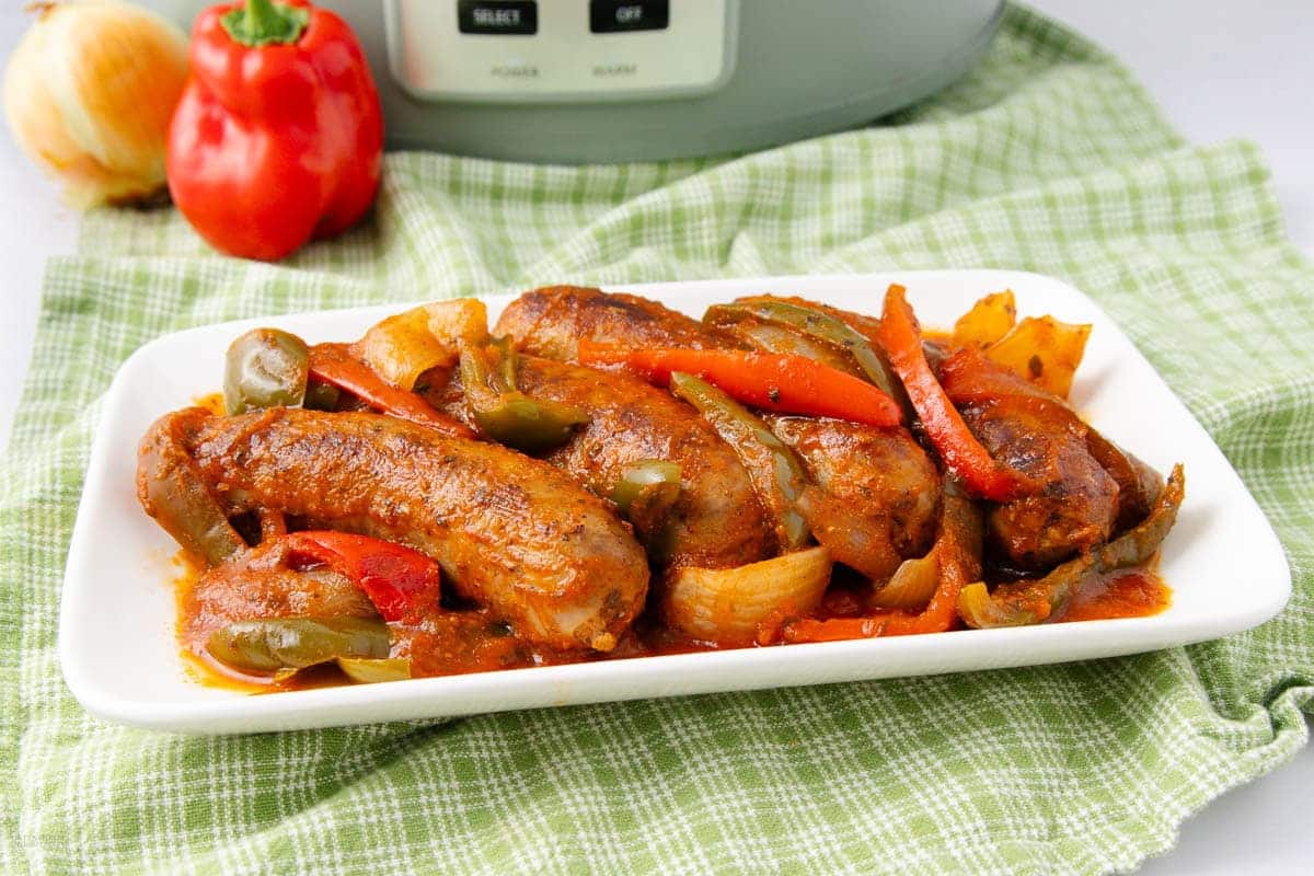 A white platter of cooked sausages with sautéed onions and bell peppers in a red sauce.