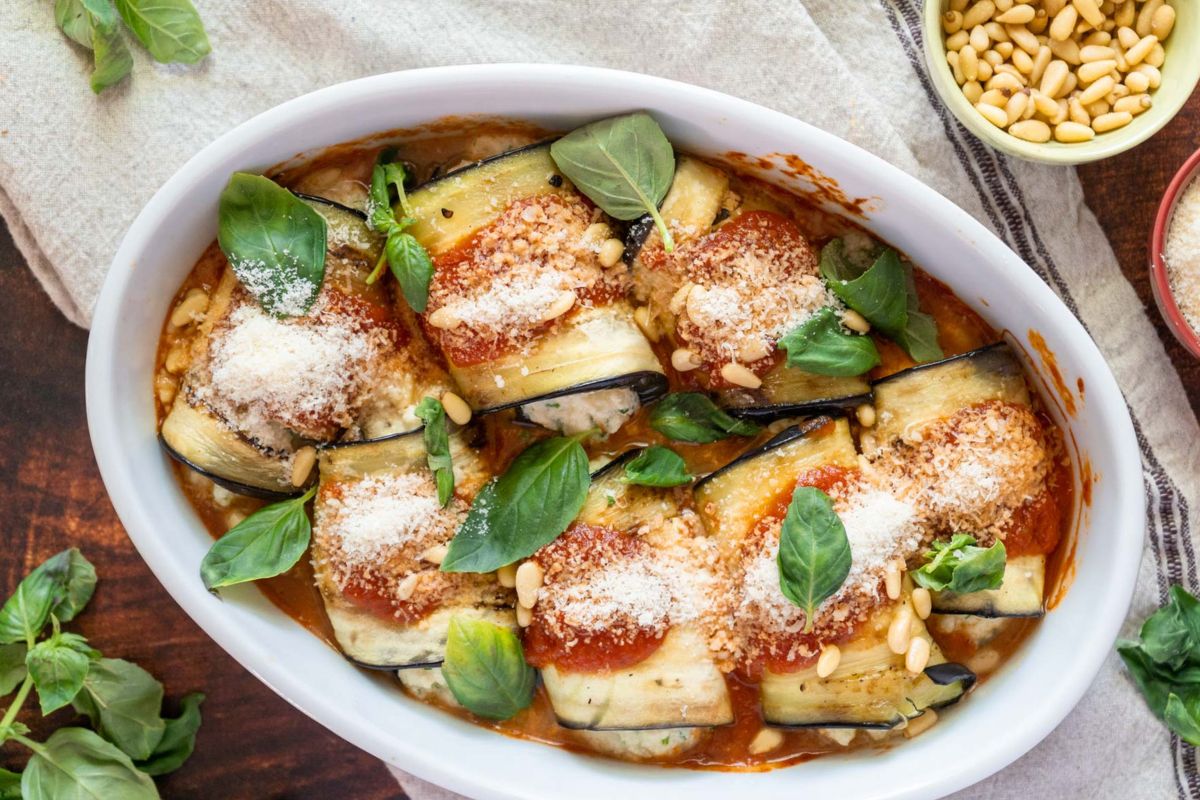 Eggplant rollatini in a baking dish topped with tomato sauce, pine nuts, parmesan, and fresh basil.