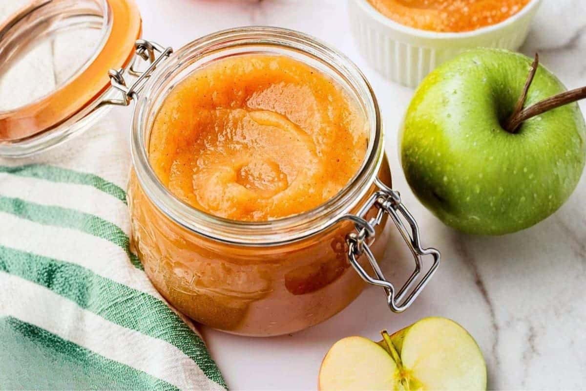 Glass jar filled with applesauce next to a whole green apple and a sliced apple.