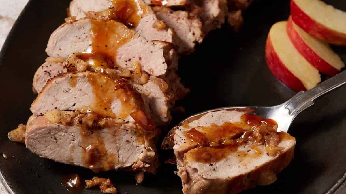 Sliced pork tenderloin with glaze on a plate next to apple slices.
