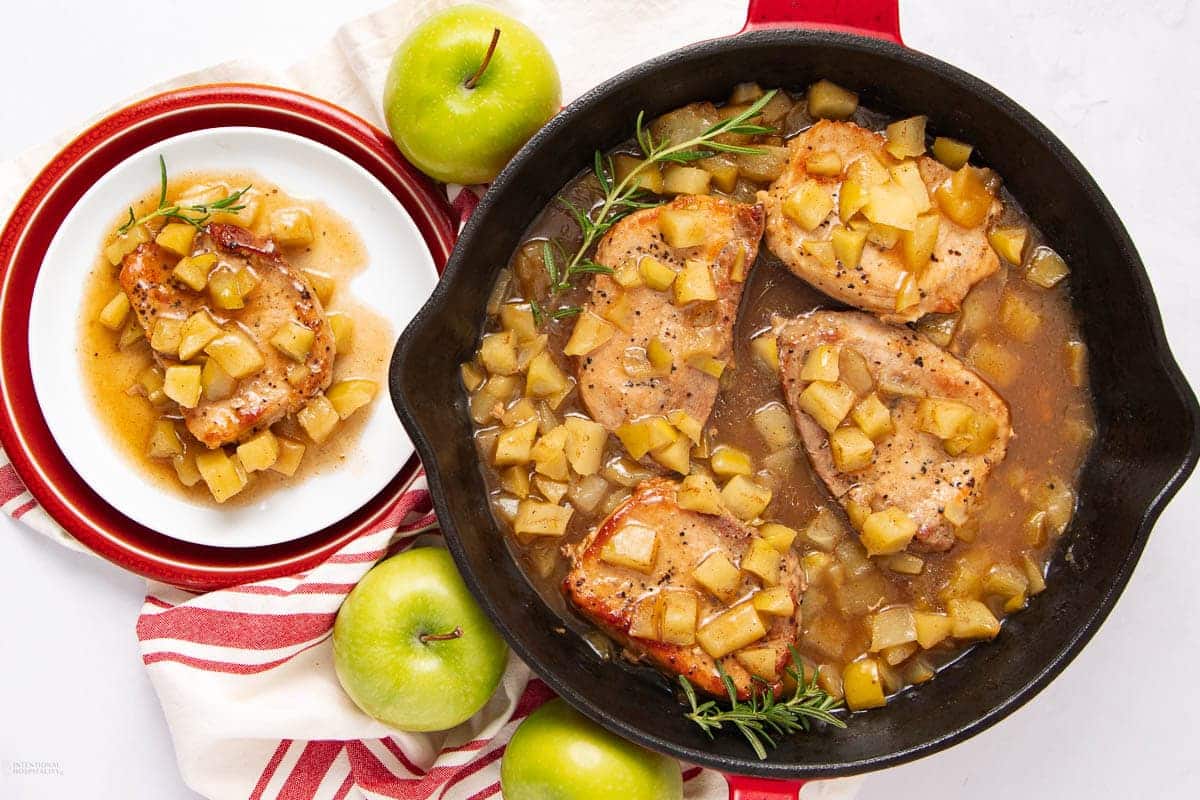 Pork chops cooked in a skillet with apples and rosemary, with one serving on a small plate.
