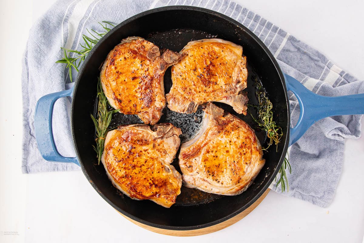 Four seared pork chops in a blue skillet with fresh herbs.
