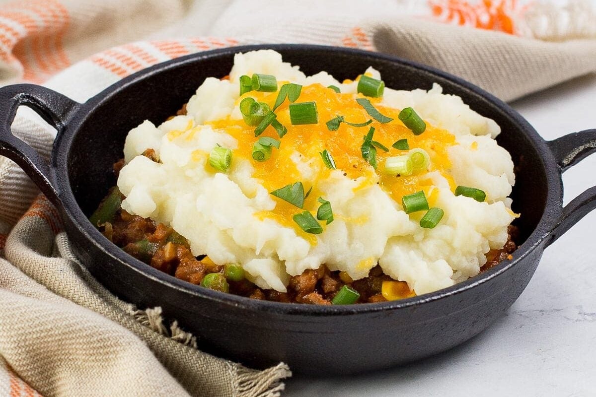 Shepherd’s pie topped with mashed potatoes, melted cheddar cheese, and green onions in a small cast iron skillet.