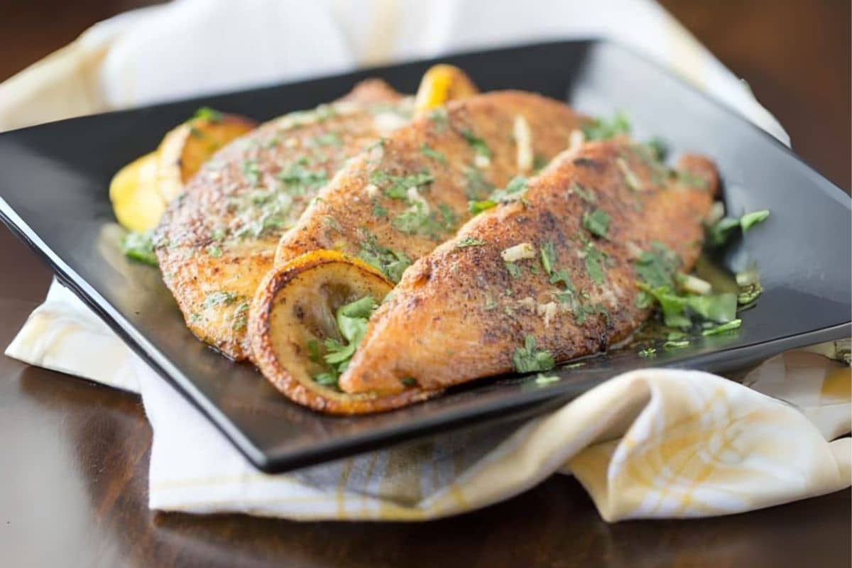 Pan-seared fish fillets topped with herbs and lemon slices on a black plate.