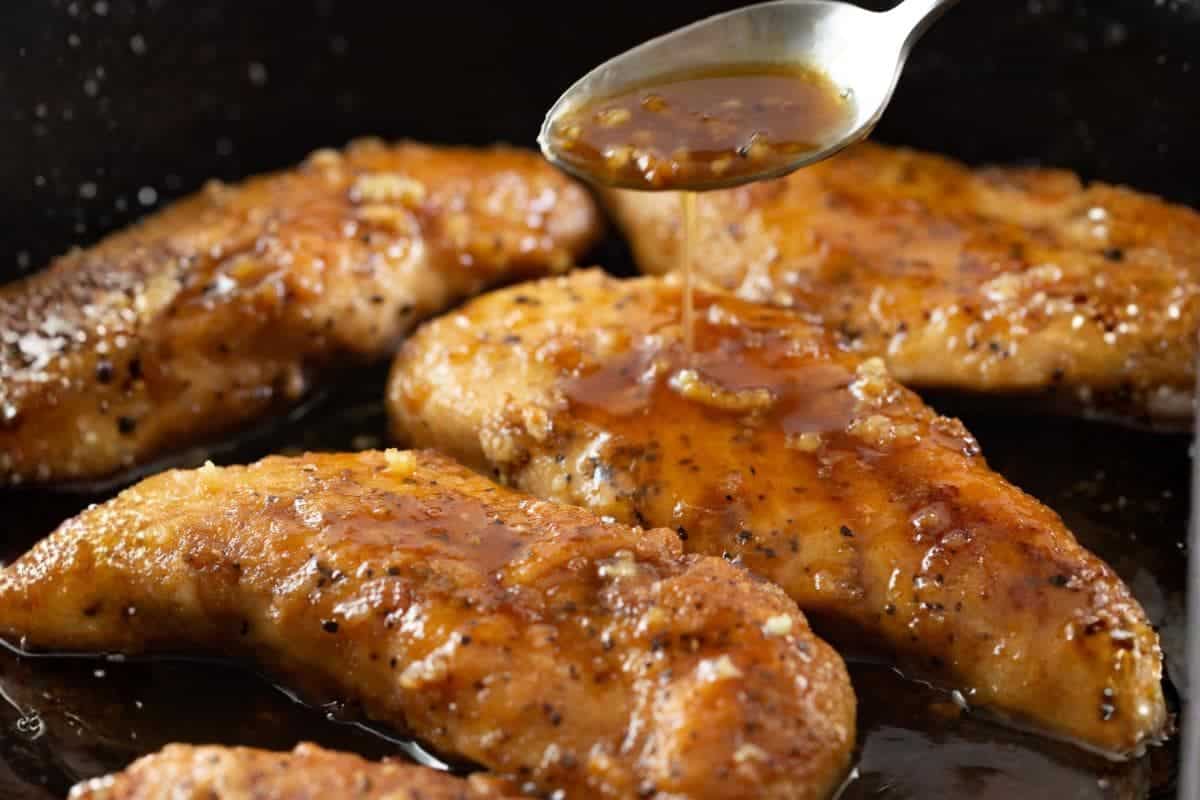 A spoon drizzling sauce over cooked chicken breasts in a pan.