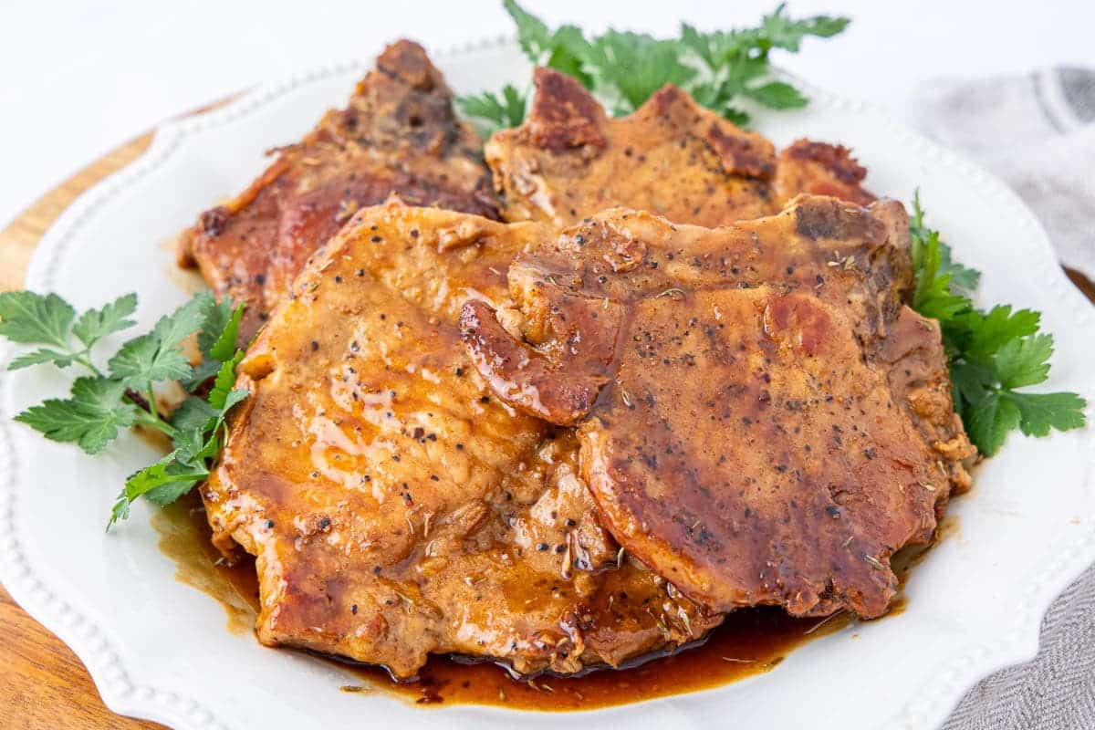 Three cooked pork chops with a dark glaze on a white plate garnished with parsley.
