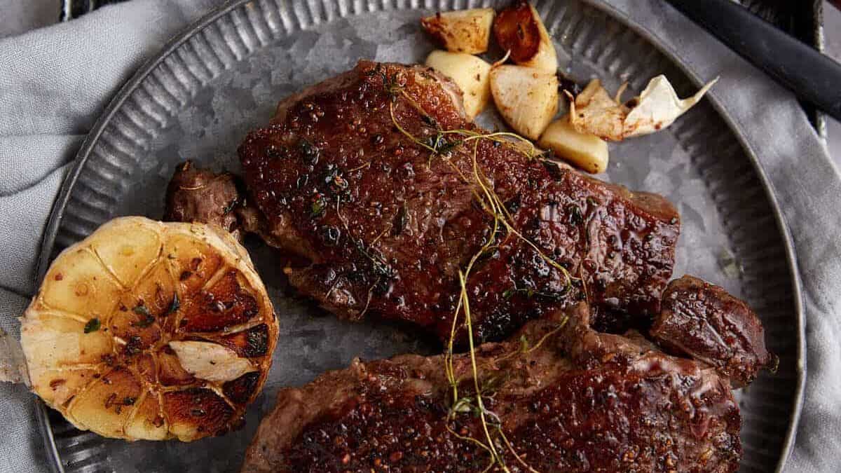 Seared steaks served with roasted garlic cloves and fresh herbs on a metal plate.