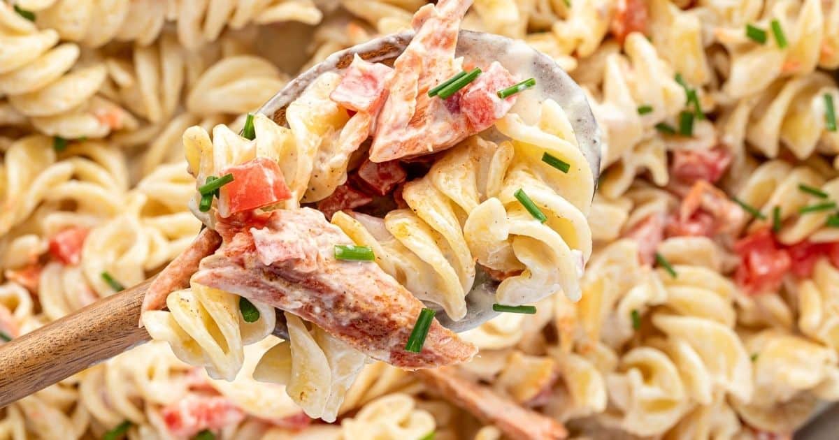 A wooden spoon lifting rotini pasta tossed in a creamy sauce with salmon, tomatoes, and chives.